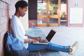 Attentive schoolgirl sitting against brick wall and using laptop