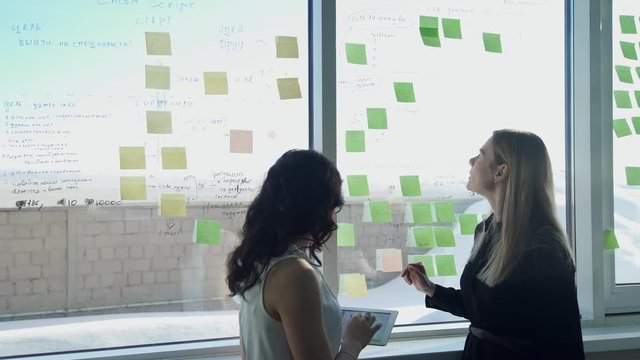 Two business women on background of window in hall discuss topics. tall blonde woman with straight hair in black shirt speaking to employee, says important ideas in office. Glass with notes, pasted