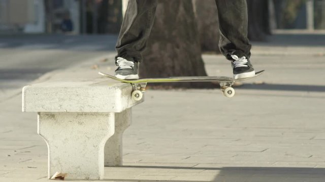 SLOW MOTION CLOSE UP DOF: Unrecognizable skateboarder jumping and sliding on concrete bench on the street. Skater's legs skateboarding and doing grinding nose slide trick with skateboard in a city