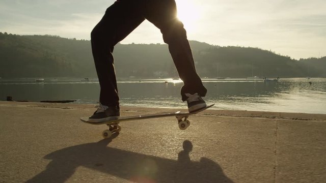 SLOW MOTION CLOSE UP Cool young skateboarder skateboarding, jumping and doing ollies on boulevard along the ocean at golden light sunset. Skateboarder riding skateboard performing nose manual trick