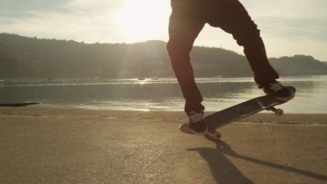 SLOW MOTION CLOSE UP Cool young skateboarder skateboarding, jumping and doing ollies on boulevard along the ocean at golden light sunrise. Skateboarder riding skateboard performing nose manual trick