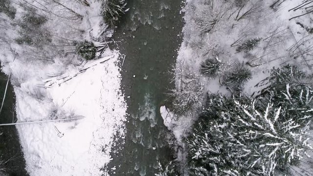 Drone Flying in Winter Storm Following Forest River Overhead with Snow Flakes Falling