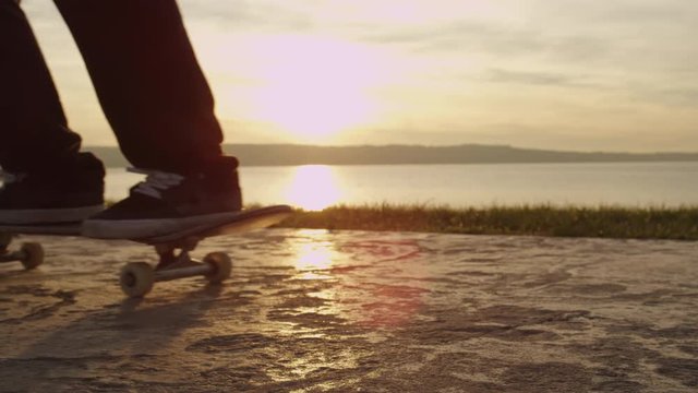 SLOW MOTION, CLOSE UP, DOF: Unrecognizable male skateboarder jump on skateboard and starts his skateboarding day at golden light sunrise. Skater skating on the concrete pavement near the ocean coast