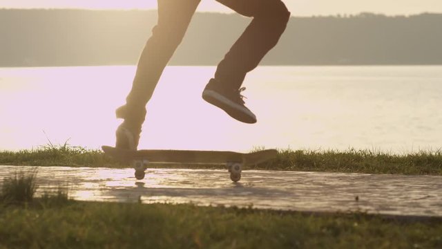 SLOW MOTION, CLOSE UP, DOF: Unrecognizable skateboarder skateboarding on the boulevard along the ocean bay at golden light sunset. Skater skating, doing spin trick on the pavement along the seashore