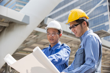 Engineer and Foreman outside office with yellow, white helmet point to building in the city with a blank plan as for construction, Teamwork Concept
