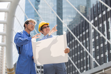 Engineer and Foreman outside office with yellow, white helmet point to building in the city with a blank plan as for construction, Teamwork Concept