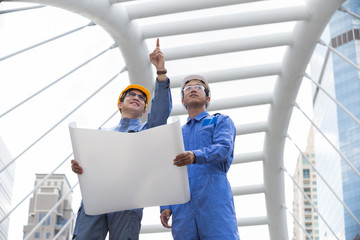 Engineer and Foreman outside office with yellow, white helmet point to building in the city with a blank plan as for construction, Teamwork Concept