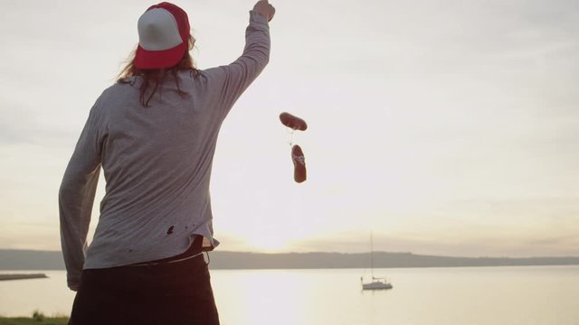 SLOW MOTION CLOSE UP, DOF: Young man with torn shirt throws sneakers away and gives up learning how to skateboard after log unsuccessful day. Angry guy with long hair throwing his shoes into the ocean