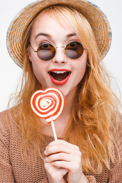 Surprised young happy blonde in sunglasses and straw hat with lollipop. Fashionable nice young hipster woman eating colorful candy, close up. Joy, fun