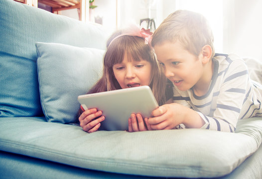 Happy siblings lying on sofa at home and playing with tablet together