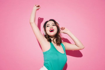 Beautiful asian girl with professional makeup and stylish hairstyle singing and dancing while listening to music on pink background.