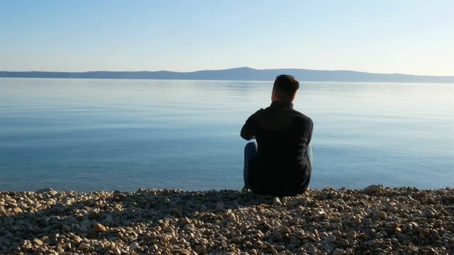 Young man sitting on the beach close to the calm blue sea and throwing pebbles