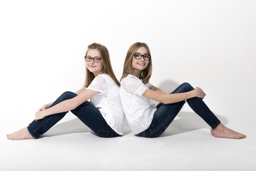 Two young girls, dressed in blue jeans and white tops, sitting back to back and smiling at the camera. The girls are isolated against a bright white background