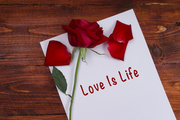 Red rose on sheet of paper with text Love Is Life on wooden table, top view.