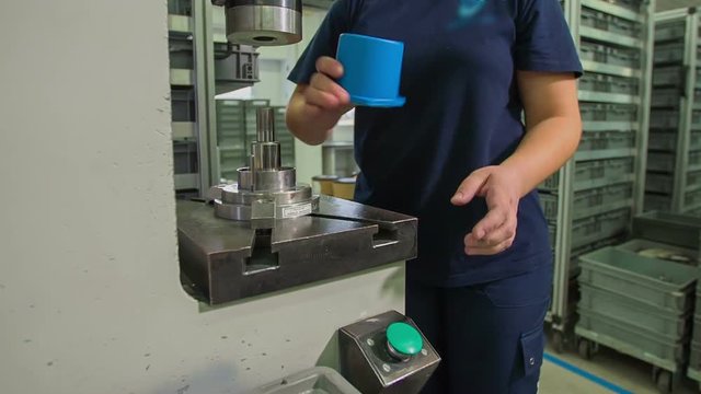 A young woman is supervising the production process of small blue items. She is then placing them inside a grey box.
