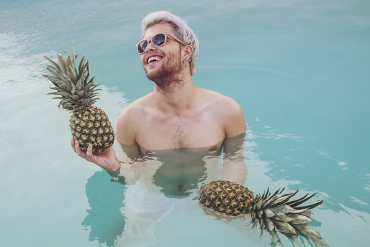Smiling young man holding pineapples in swimming pool