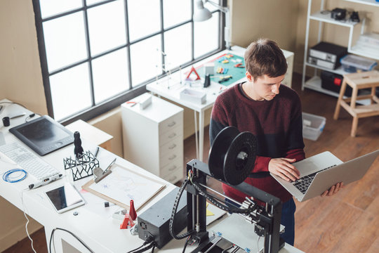 Young Caucasian man graphic setting up 3d printer on his laptop.