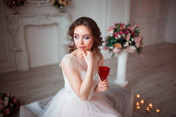 Girl with makeup in a pink wedding dress is sitting in a beautiful room surrounded by flowers and candles. The bride holds in hands red wine glass
