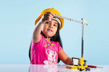 cute indian baby girl playing with toy crane wearing yellow construction hat or hard hat, childhood and education concept