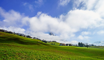 Alpine natural landscape with green fields, high rocks and white clouds