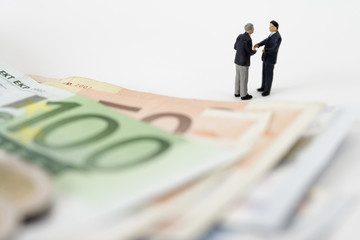 Two businessmen figurines shaking hands, Euro banknotes in foreground