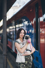 Beautiful young woman holding her pug dog in arms and standing on train station.