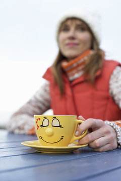 Young woman in warm clothing sitting outside at a table with a painted cup in her hand, selective focus