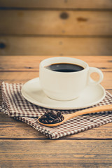 Coffee beans in spoon with a cup of coffee on wood table.