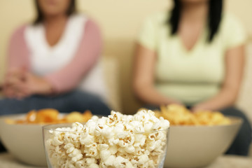 Two full figured women sitting behind three bowls filled with popcorn and crisps, selective focus