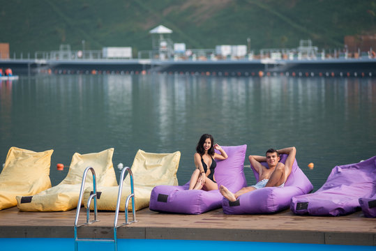 Young sexy couple on cushioned loungers by swimming pool and lake on the background. Attractive brunette girl is wearing a black swimsuit