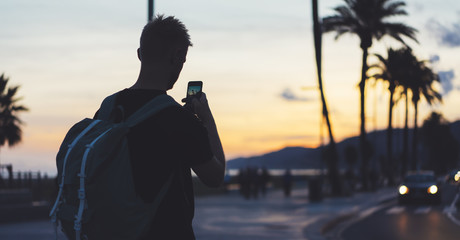 Fototapeta premium Man with backpack making photo sunset beach on smart phone, tourist look and enjoy nature on background color sky and taxi on palm summer street, hipster using in hands mobile phone, traveler mock up