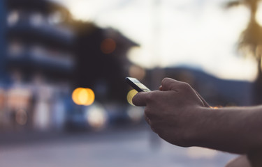 Man pointing finger on screen smart phone on background sunset color light in night atmospheric city, hipster using in male hands and text mobile phone, mock up palm street, content relax lifestyle