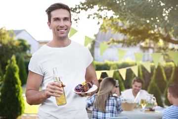 Young man holding bottle of beer and plate with grilled meat and vegetables