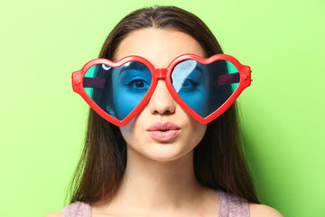 Beautiful young woman with heart-shaped glasses on color background