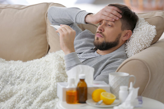 Young ill man with electronic thermometer lying on sofa at home