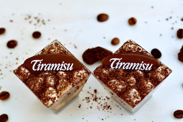 Top view of two tiramisu glasses on coffee beans, cocoa powder, chocolate pieces background closeup - traditional Italian dessert
