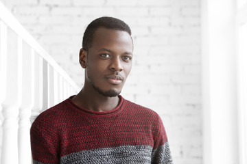 Attractive young African American male dressed casually looking at camera with dreamy expression, deep in thoughts, standing alone in white room with brick walls, leaning his shoulder on columns