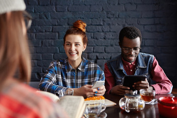 Staying connected. Modern young interracial couple using electronic gadgets, surfing internet and enjoying high-speed wireless connection while having lunch with unrecognizable girl with book