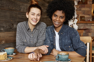 Sweet shot of happy beautiful Caucasian redhead woman holding hands with her stylish African American girlfriend while having breakfast at modern coffee shop, sitting at table with mugs and cake