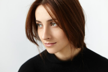 Naklejka premium Studio shot of attractive European model with dark hair having dreamy and thoughtful expression on her face, looking sideways with faint smile, posing against white wall background wearing black dress