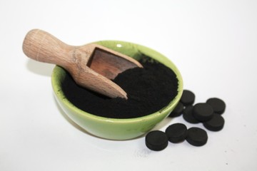 Black activated charcoal powder in a bowl with wooden utensil and few pills next