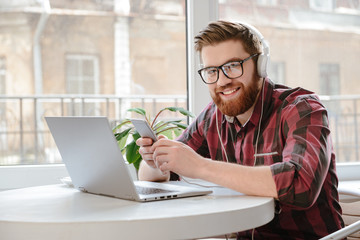Cheerful young man using phone and laptop while listening music.