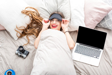 Top view on the young woman lying with eyesight on the bed with laptop, phone and photo camera. Resting after the work