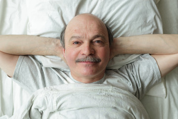 An elderly man slyly lies on the bed smiling. Top-down view