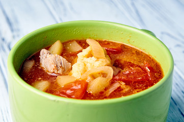 A bowl of soup close-up. A green tureen on a white wooden table. Thick red soup with meat and dumplings. A traditional dish of Hungarian cuisine is Goulash. 