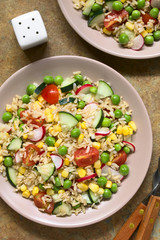 Brown rice salad with cherry tomato, corn, cucumber, radish and peas served on plate, photographed overhead on slate with natural light