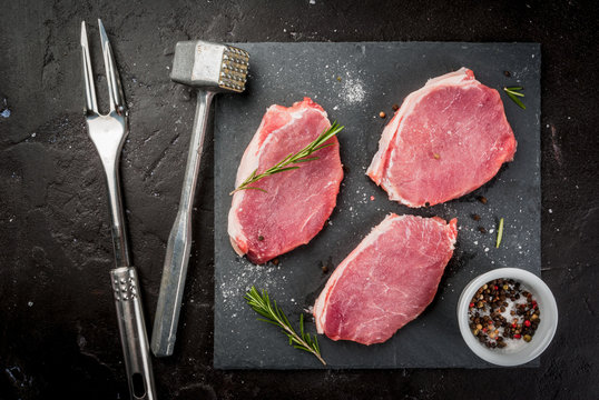 Fresh raw pork, steaks, on a cutting shale board on a black concrete table. Top view, with a hammer to beat the meat and a fork, copy space