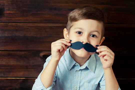 A kid with props for a photo booth. A child with the requisite mustache on wooden background.