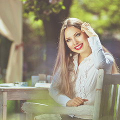 Beautiful colorful toned portrait of young woman with a long hair, elegantly dressed in white shirt, sitting on wooden chair at  sunny spring day with a tree on background