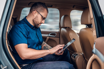 Obraz premium A man in eyeglasses with tattoo on his arm using portable tablet PC on a back seat of a car.